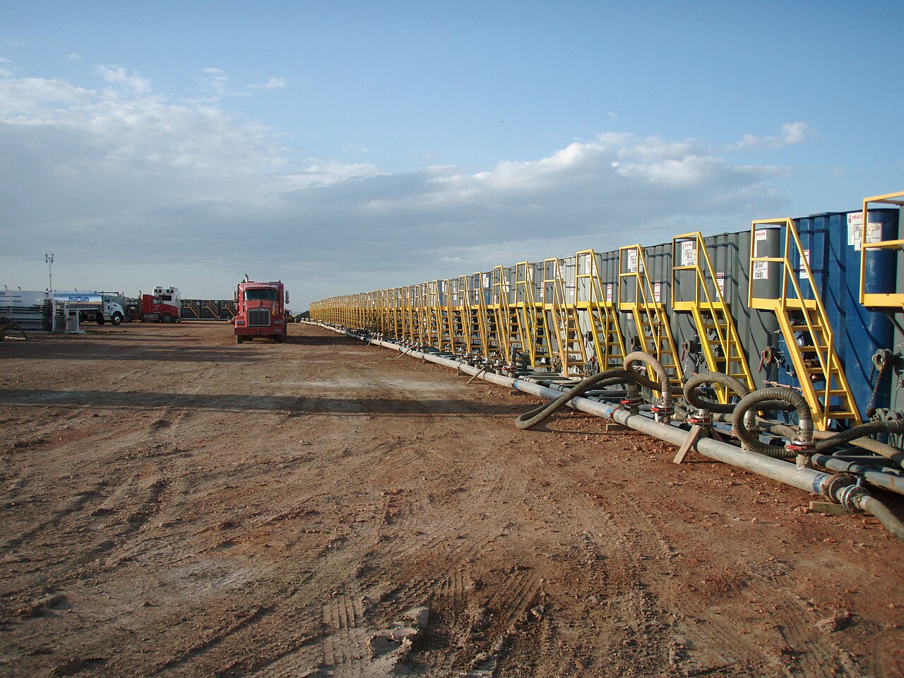 A long row of blue portable steel tanks with yellow access stairs, connected by hoses to a pipe manifold on bare ground, with trucks in the distance